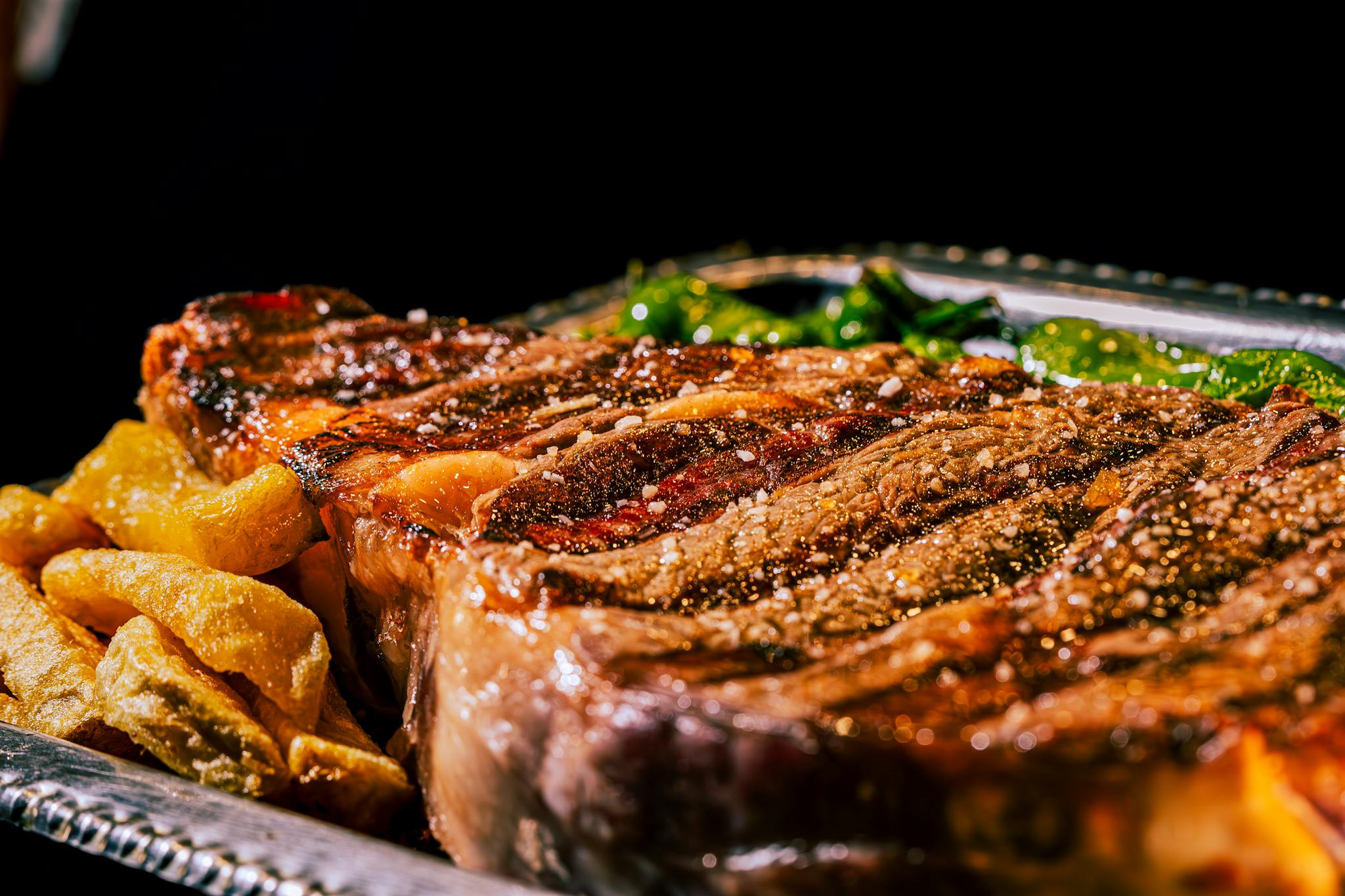 Close-up of a juicy grilled ribeye steak with pepper and sides on a silver platter.